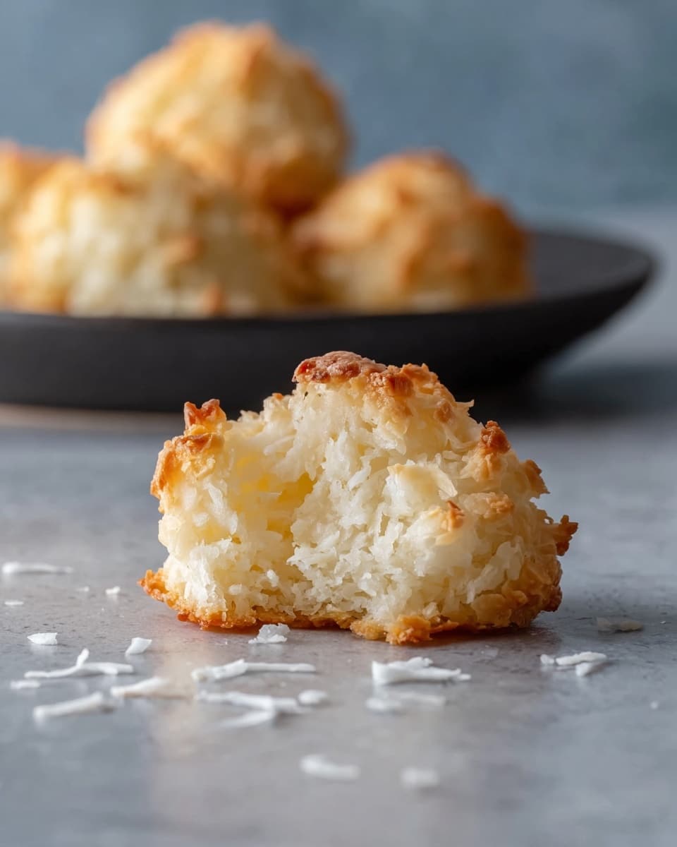 The image showcases a close-up of a freshly baked coconut macaroon, featuring a golden-brown crust with a soft, chewy interior made up of shredded coconut. A few coconut flakes are scattered on the smooth surface beneath it, enhancing the texture and inviting a taste. In the blurred background, additional macaroons sit on a dark plate, suggesting a delightful batch ready to be enjoyed. The neutral tones of the background highlight the warm colors of the macaroon, making it the focal point of the image. photo taken with an iPhone --ar 4:5 --v 7