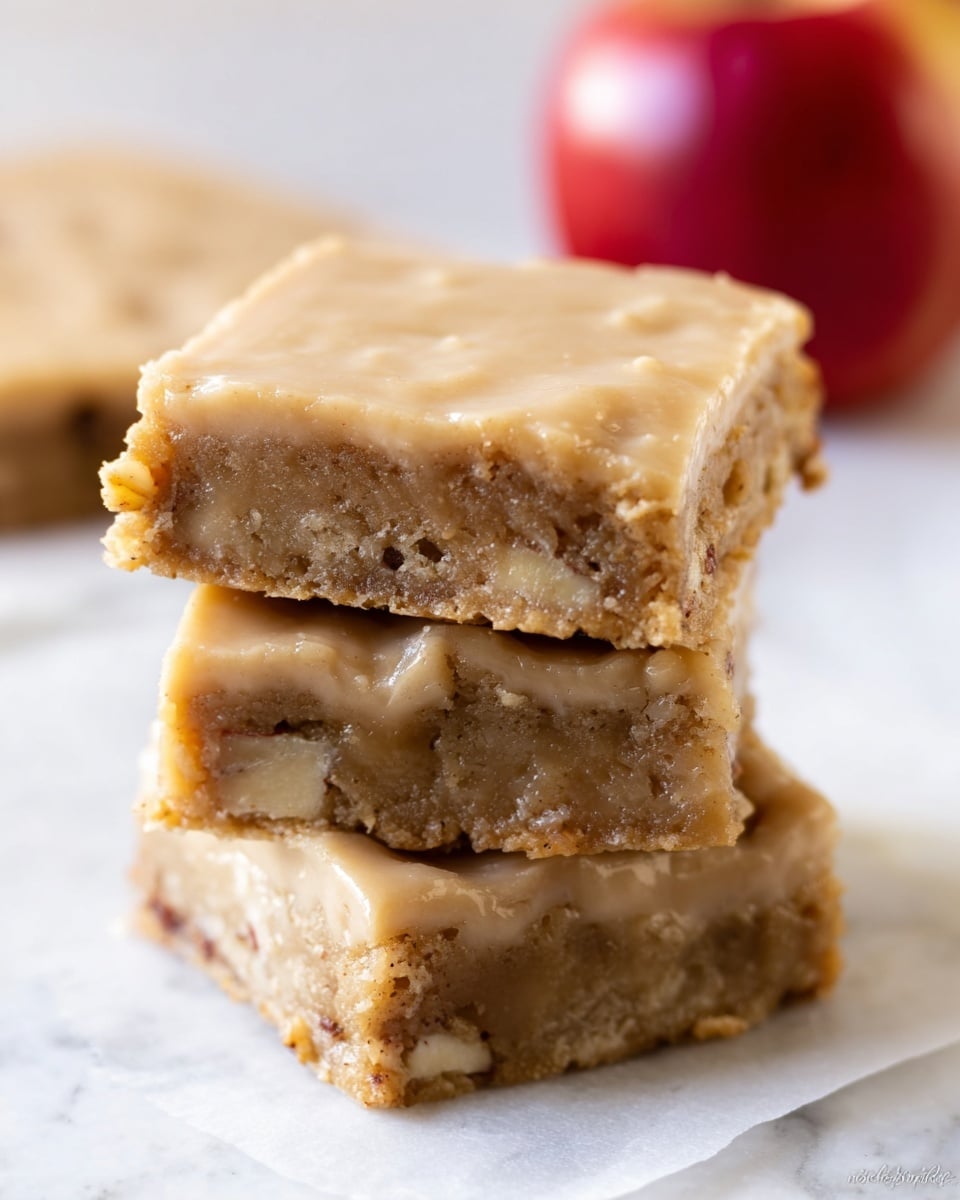 The image shows a stack of three square dessert bars placed on a white marbled surface, each bar having two main layers. The bottom layer is a dense, crumbly light brown crust with small visible chunks of what appears to be nuts or fruit inside. The top layer is a smooth, glossy light tan glaze that evenly coats the surface. The bars look thick and moist, with a soft texture visible on the sides. In the blurred background, a red apple is visible, adding context to the flavor of the dessert. photo taken with an iphone --ar 4:5 --v 7