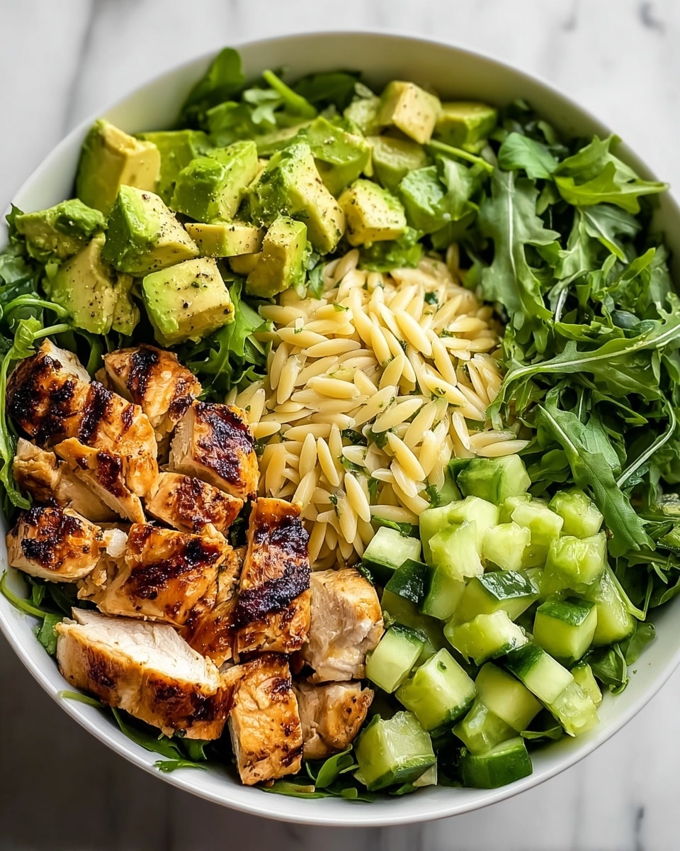 A white bowl filled with four main layers starting from the base with fresh green leafy arugula covering the bottom. On top of the arugula, the bowl is divided into four sections: bright green chopped avocado with a slightly creamy texture and black pepper sprinkled on it; golden-brown grilled chicken pieces with visible grill marks showing a slightly charred texture; light yellow orzo pasta garnished with herbs, appearing soft and tender; and small cubes of fresh, bright green cucumber with a moist look. The ingredients are neatly arranged so each section is clearly separated, and the photo has a clean look with a white marbled surface beneath the bowl. Photo taken with an iphone --ar 4:5 --v 7