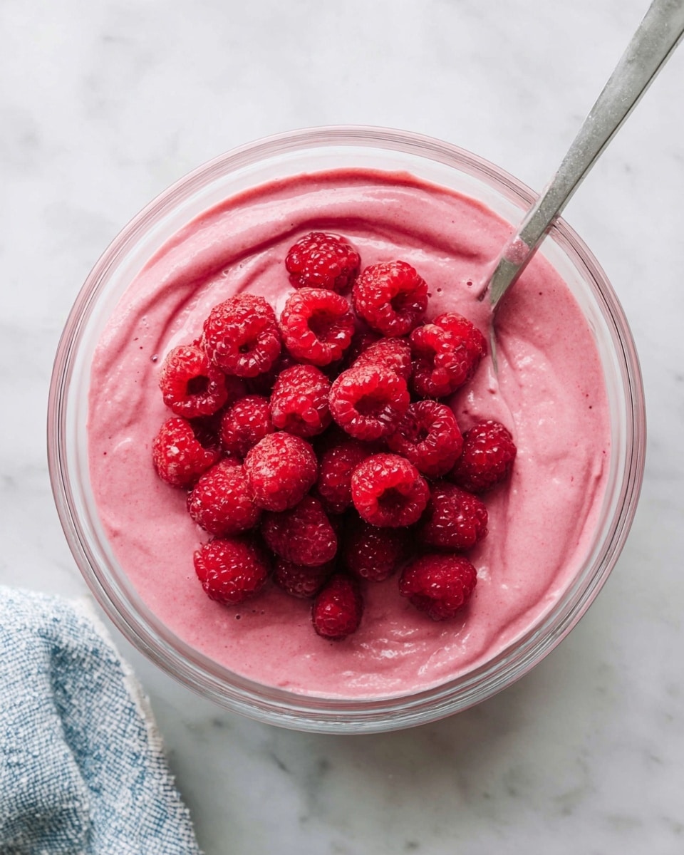 A clear bowl filled with a thick, smooth pink mixture, topped with a pile of whole, bright red raspberries clustered in the center. A silver spoon is resting inside the bowl, partially dipped in the pink mixture on one side. The bowl is placed on a white marbled surface with part of a blue and white cloth visible at the bottom left corner. photo taken with an iphone --ar 4:5 --v 7