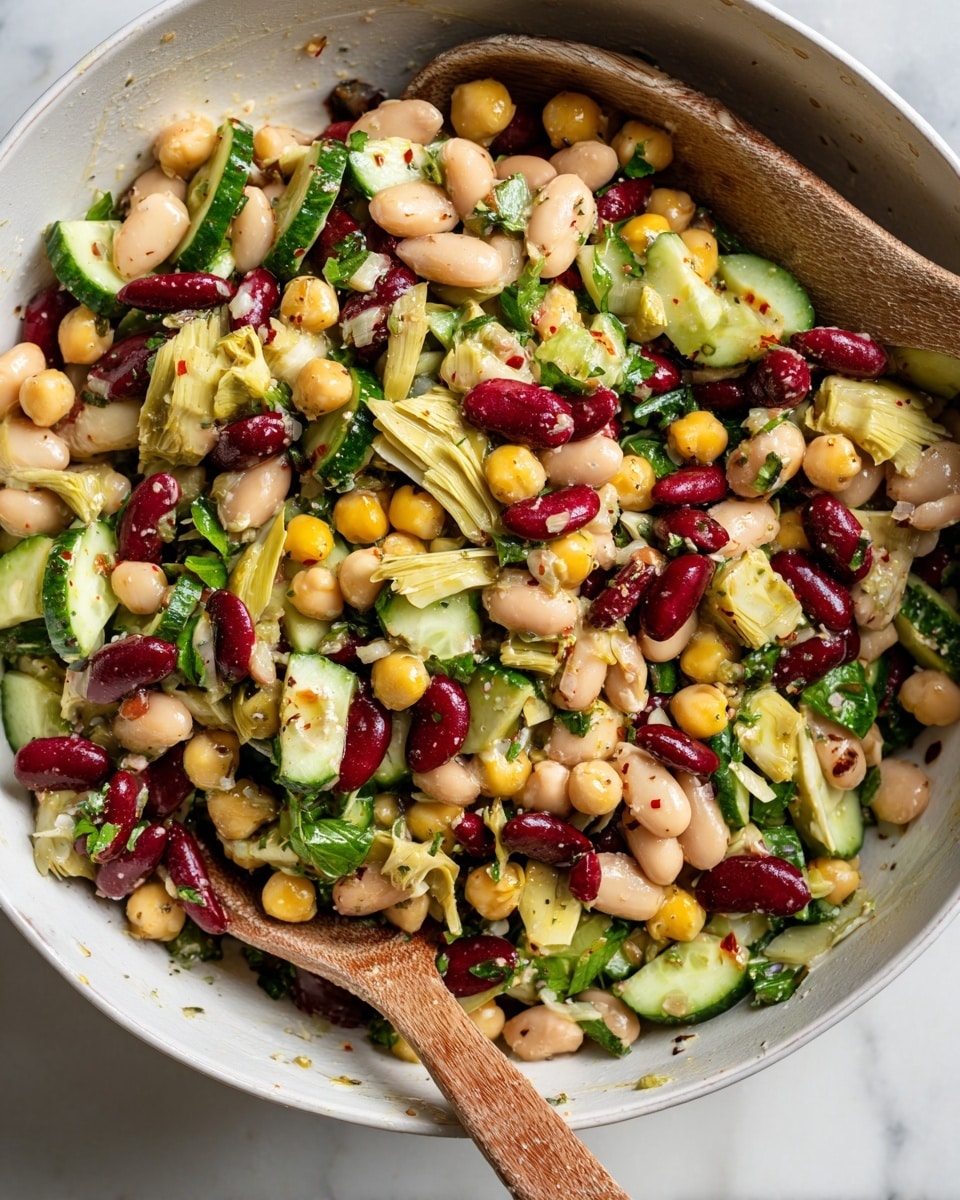 A white speckled bowl is filled with a colorful beans salad featuring three layers of different beans: pale chickpeas, white beans, and dark red kidney beans. Mixed throughout are green cucumber slices, pale yellow artichoke hearts, and sun-dried tomato pieces, adding red and tan tones along with fresh green herbs scattered on top. The bowl sits stacked on a solid white bowl, which rests on a matte dark gray plate, all placed on a white marbled surface. A silver fork is placed inside the bowl resting on the salad, and in the background, there is a white jar with oil and a white cloth with beige stripes. A sprig of fresh parsley is seen at the bottom edge of the image. photo taken with an iphone --ar 4:5 --v 7