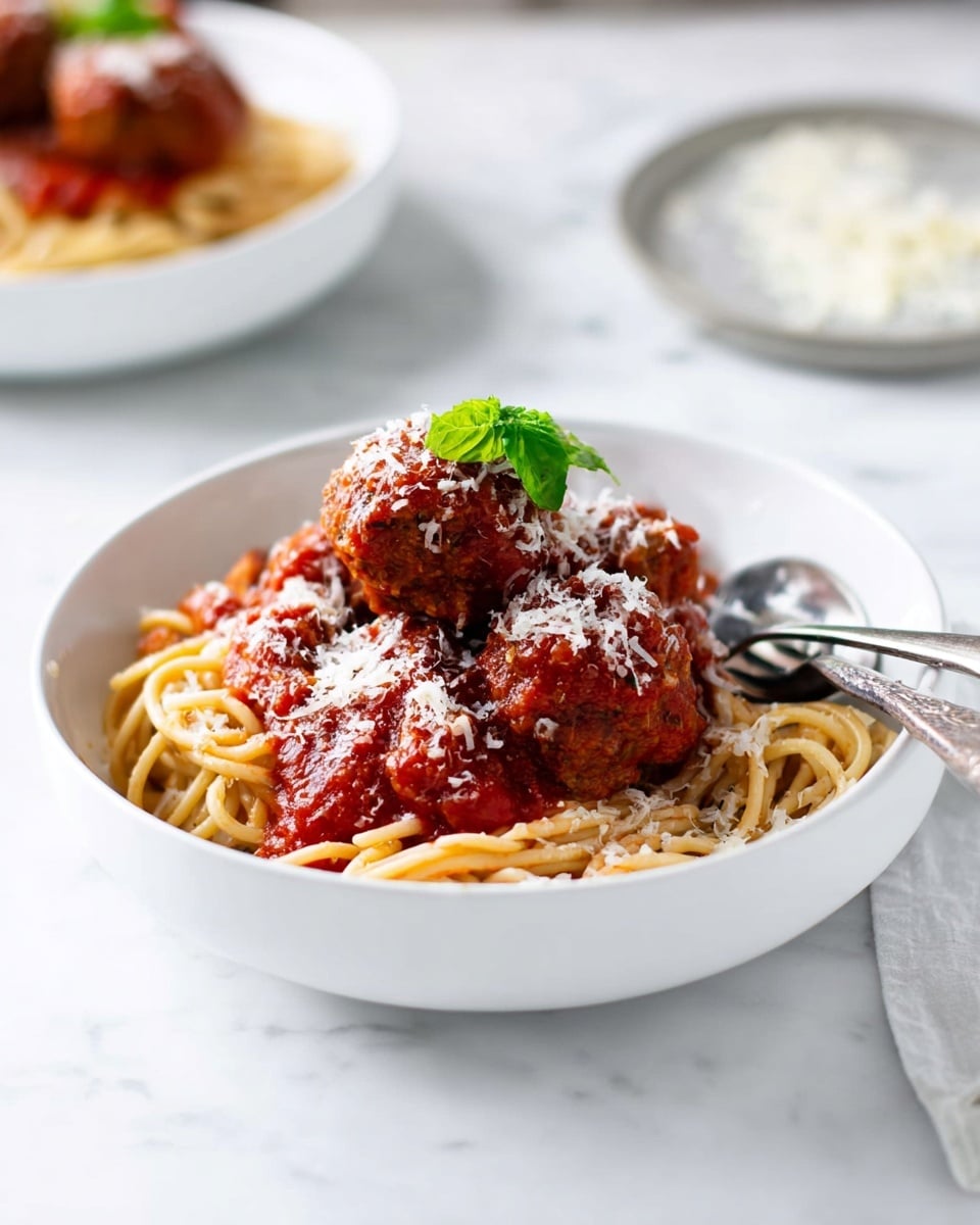 A white bowl holds a neat nest of light brown spaghetti as the base layer, topped with several round, rich brown meatballs covered in thick red tomato sauce, which also spreads slightly over the pasta. White grated cheese is sprinkled over the meatballs and sauce, adding a fine texture, and a small sprig of bright green basil rests on top for color contrast. A silver fork and spoon lie inside the bowl on the right side. The dish sits on a white marbled surface with another similar plate blurred in the background. Photo taken with an iphone --ar 4:5 --v 7