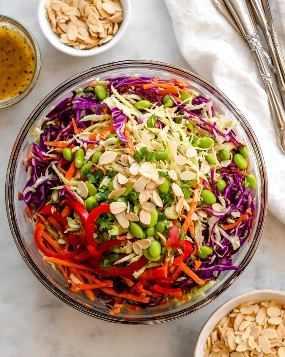 The image shows a clear glass bowl filled with a colorful salad that has many layers of thinly sliced vegetables and nuts. The base layer consists of shredded purple and white cabbage mixed with bright orange carrot strips and green edamame beans. On top, there are sliced red bell peppers, pale toasted almond slices, and green chopped herbs adding texture and color. The bowl is on a white marbled surface, with a small bowl of dressing in the top left, and a small bowl of almond slices on the bottom right. In the upper right corner, there is a white cloth with silver serving utensils. photo taken with an iphone --ar 4:5 --v 7