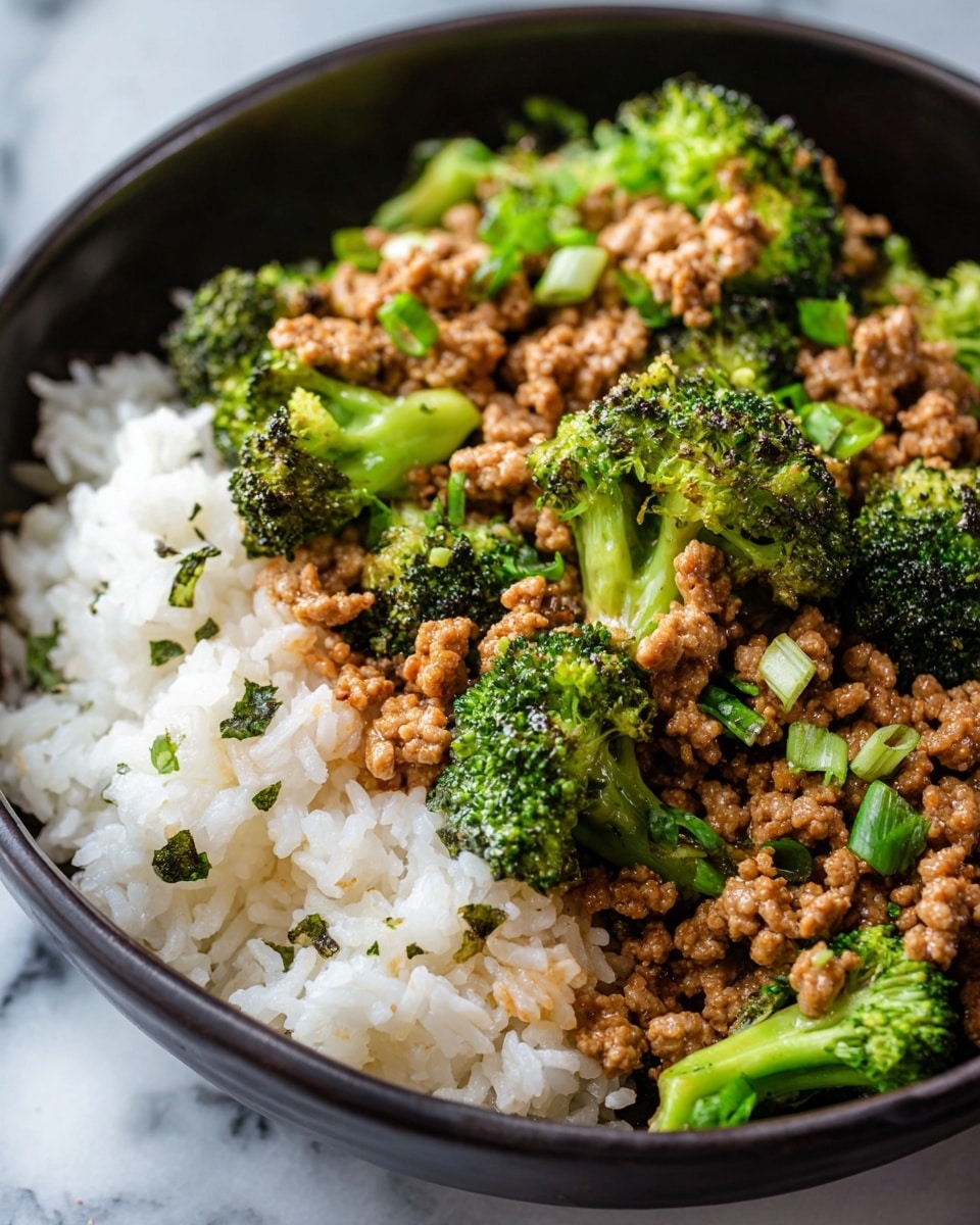 A close-up view of a dark bowl filled with two main layers: the bottom layer is fluffy white rice with a soft texture, lightly scattered with bits of green herbs, occupying about half of the bowl. On top, covering the other half and slightly overlapping the rice, there is a mix of cooked ground meat and bright green broccoli florets. The ground meat looks moist and seasoned, coated with a light brown sauce that adds shine. The broccoli pieces are vivid green, with some showing a slightly charred texture. Small pieces of chopped green onions are sprinkled on top, giving contrast and freshness. The bowl sits on a white marbled surface, photo taken with an iphone --ar 4:5 --v 7
