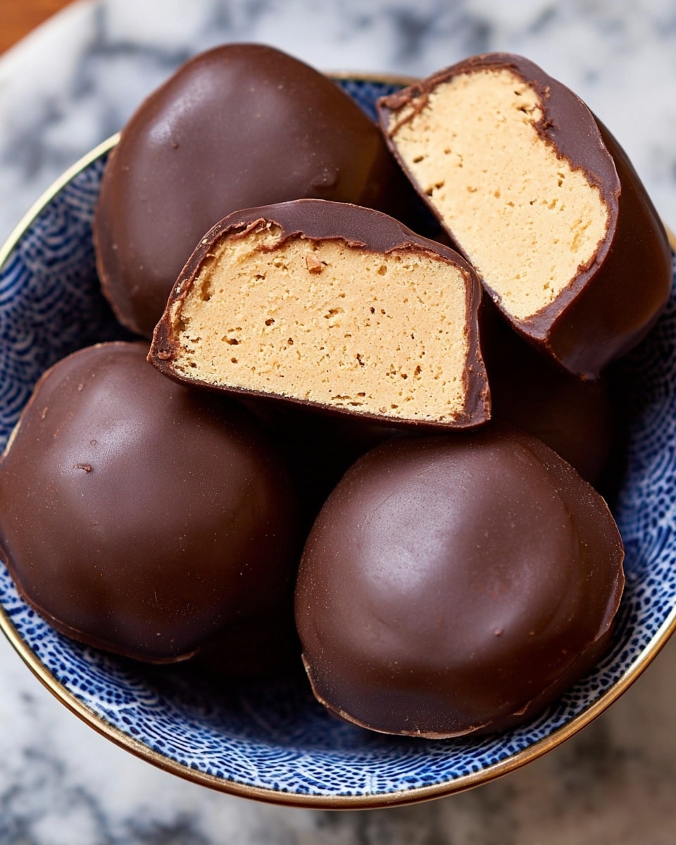A close-up top view of five round chocolate-covered treats placed inside a white bowl with a blue pattern, on a white marbled surface. Three whole treats appear smooth and shiny, colored dark brown on the outside, and two are cut in half, showing a thick layer of soft, light tan filling with a slightly porous texture inside, surrounded by a thin dark brown chocolate shell layer. The halves are positioned on top of the whole ones, showing the contrast between the smooth outer chocolate and the soft inner filling. Photo taken with an iphone --ar 4:5 --v 7