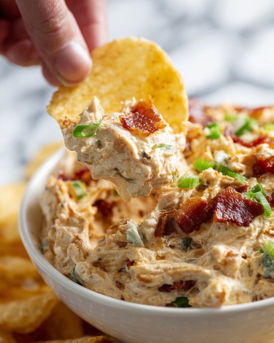 The image shows a close-up of a potato chip dipped into a creamy dip in a white bowl. The dip has a thick, chunky texture with a light beige color and visible pieces of green onions and crispy bacon mixed throughout. The bowl is filled with the dip, and a woman's hand is holding the chip, lifting it from the bowl. The background is blurred with a white marbled texture. photo taken with an iphone --ar 4:5 --v 7