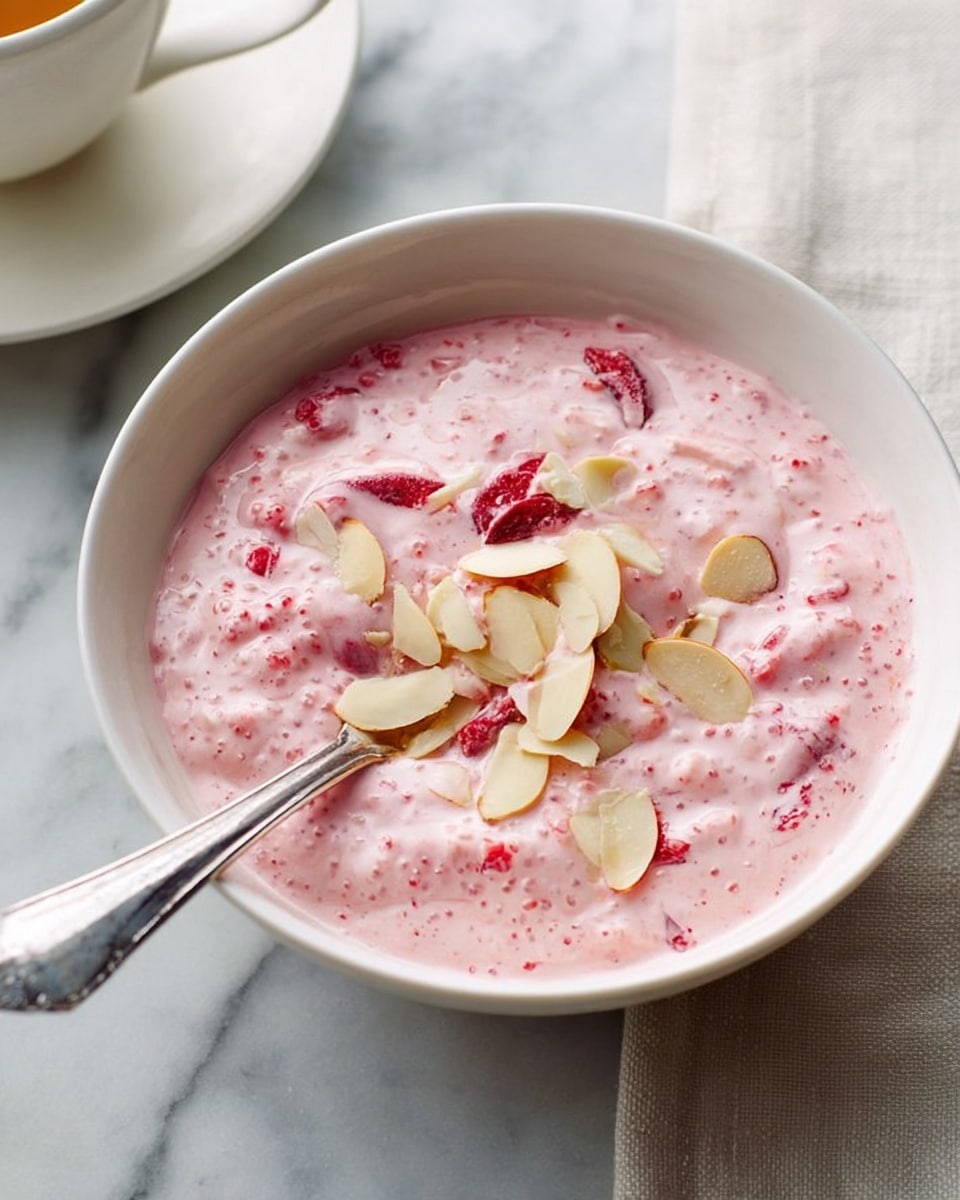 The image shows a white bowl filled with a pink creamy mixture that has visible small bubbly textures and pieces of red fruit mixed in. On top, there are scattered thin almond slices lightly spread out. A silver spoon rests inside the bowl with its handle leaning out, positioned on a white marbled surface. In the background, part of a white cup and saucer is visible near the bowl. photo taken with an iphone --ar 4:5 --v 7