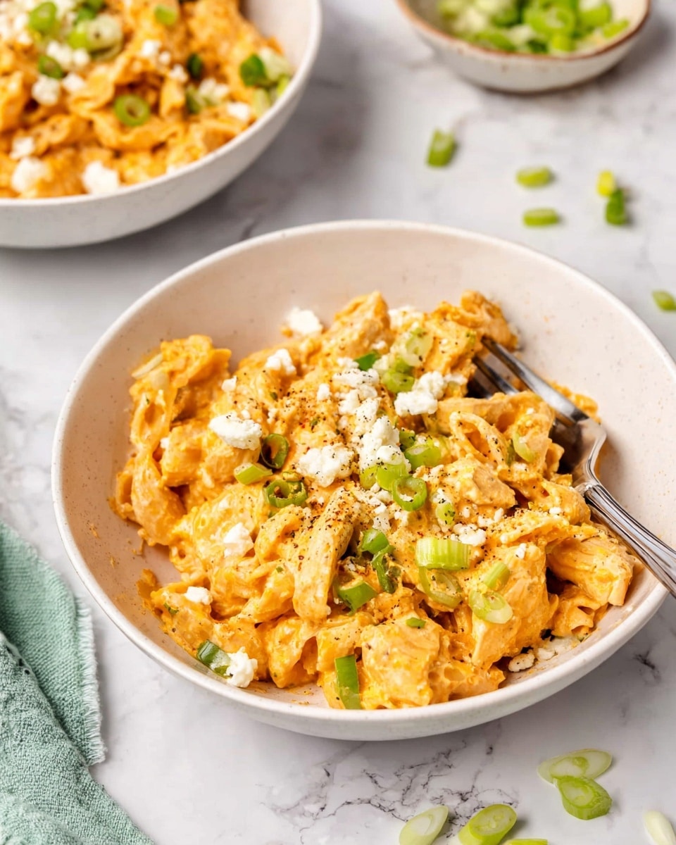 A white bowl is filled with a creamy, cheesy orange pasta mixed with chunks of white chicken and small pieces of green celery or pepper. The pasta looks soft and rich, covered in a thick, smooth cheddar sauce. On top, there are small crumbles of white cheese and thin slices of green onion scattered evenly, adding a fresh touch. A metal fork rests inside the bowl, partially buried in the pasta. The bowl sits on a white marbled surface with a few green onion pieces scattered nearby, with another bowl of the same dish blurred in the background. photo taken with an iphone --ar 4:5 --v 7