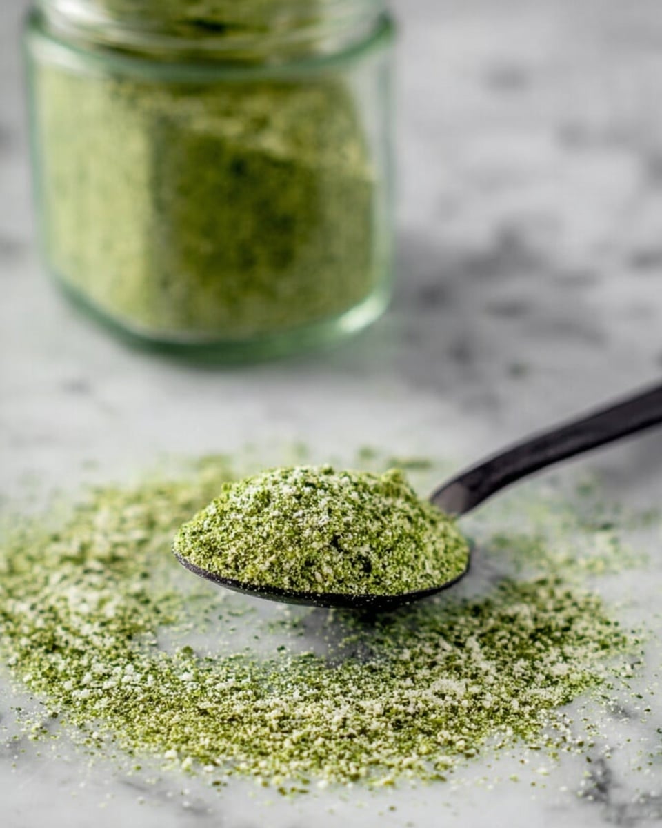 The image shows a close-up of a black spoon filled with finely ground green seasoning with small white specks, resting on a white marbled surface, surrounded by a scattered pile of the same green and white powder. In the blurred background, there is a glass jar filled with more of the green seasoning. The texture looks crumbly and slightly coarse, giving a fresh and natural feel. Photo taken with an iphone --ar 4:5 --v 7