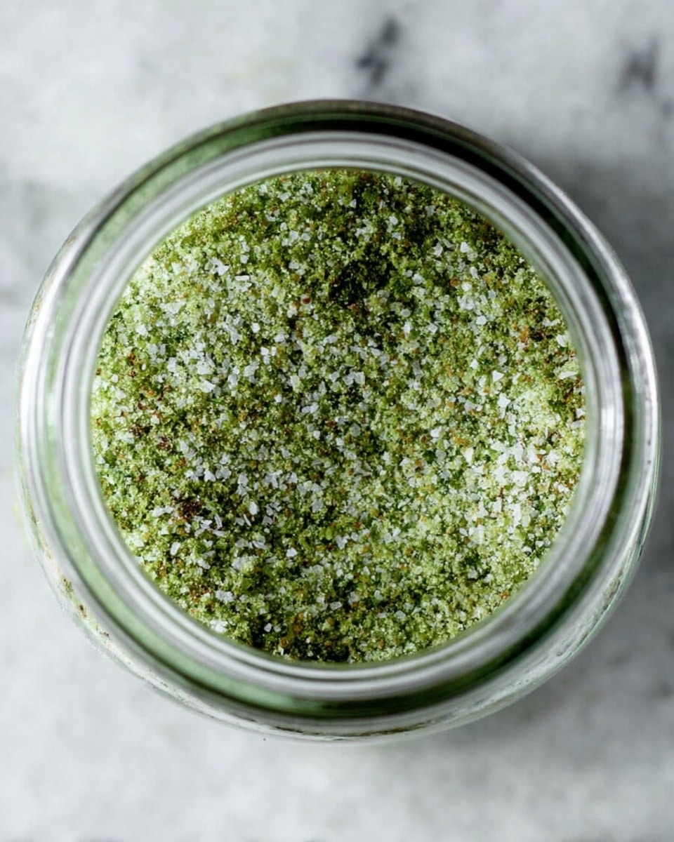A close-up view of a clear glass jar filled to the brim with a coarse green seasoning mix, showing varied textures with small white salt crystals and darker green herb flakes evenly spread across the surface. The jar is placed on a white marbled textured surface, with the round shape of the jar opening clearly visible from above, highlighting the mixture's granular details and color contrasts. photo taken with an iphone --ar 4:5 --v 7