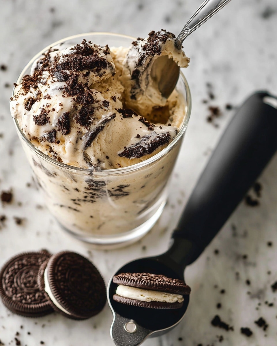 A close-up of a clear glass filled with one thick layer of light brown creamy dessert mixed with small, dark chocolate cookie pieces creating a speckled texture. A metal spoon rests on the rim of the glass, scooping a bite that shows the creamy, slightly grainy texture with chunks of cookies clearly visible. The scene is set on a white marbled surface with blurred background elements in soft neutral colors. The lighting is natural, highlighting the smooth and chunky texture of the dessert. Photo taken with an iphone --ar 4:5 --v 7