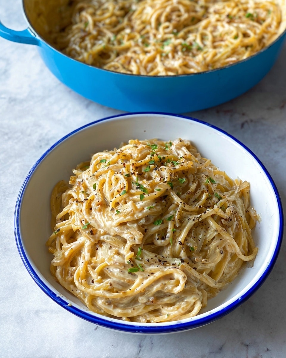 A white bowl with a thin blue rim is filled with a creamy spaghetti dish. The noodles are coated in a thick light beige sauce mixed with small pieces of cooked onions, visible ground black pepper, and bits of green herbs scattered on top. In the background, there is a blue pot containing more of the same pasta, showing a glossy, creamy texture with similar ingredients. The scene is set on a white marbled surface. Photo taken with an iphone --ar 4:5 --v 7