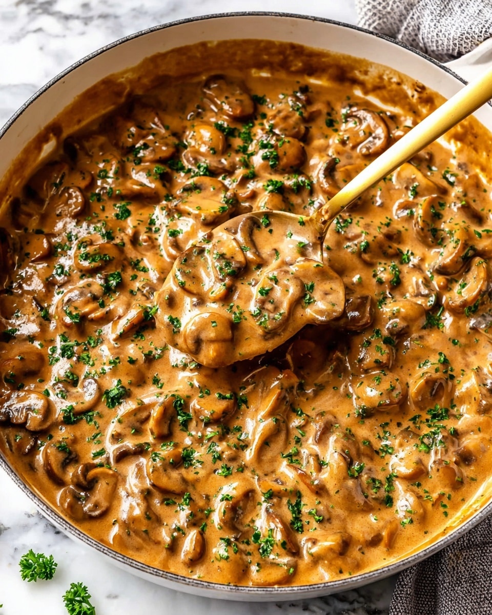 The image shows a close-up of a white pan filled with a rich, creamy mushroom sauce that has a glossy light brown color. The sauce contains many sliced mushrooms, all cooked to a tender texture, scattered evenly throughout. Bright green chopped parsley is sprinkled on top, adding a fresh pop of color. A golden spoon is partly dipped into the sauce, lifting some of the thick, smooth mixture. The pan is placed on a white marbled surface. photo taken with an iphone --ar 4:5 --v 7
