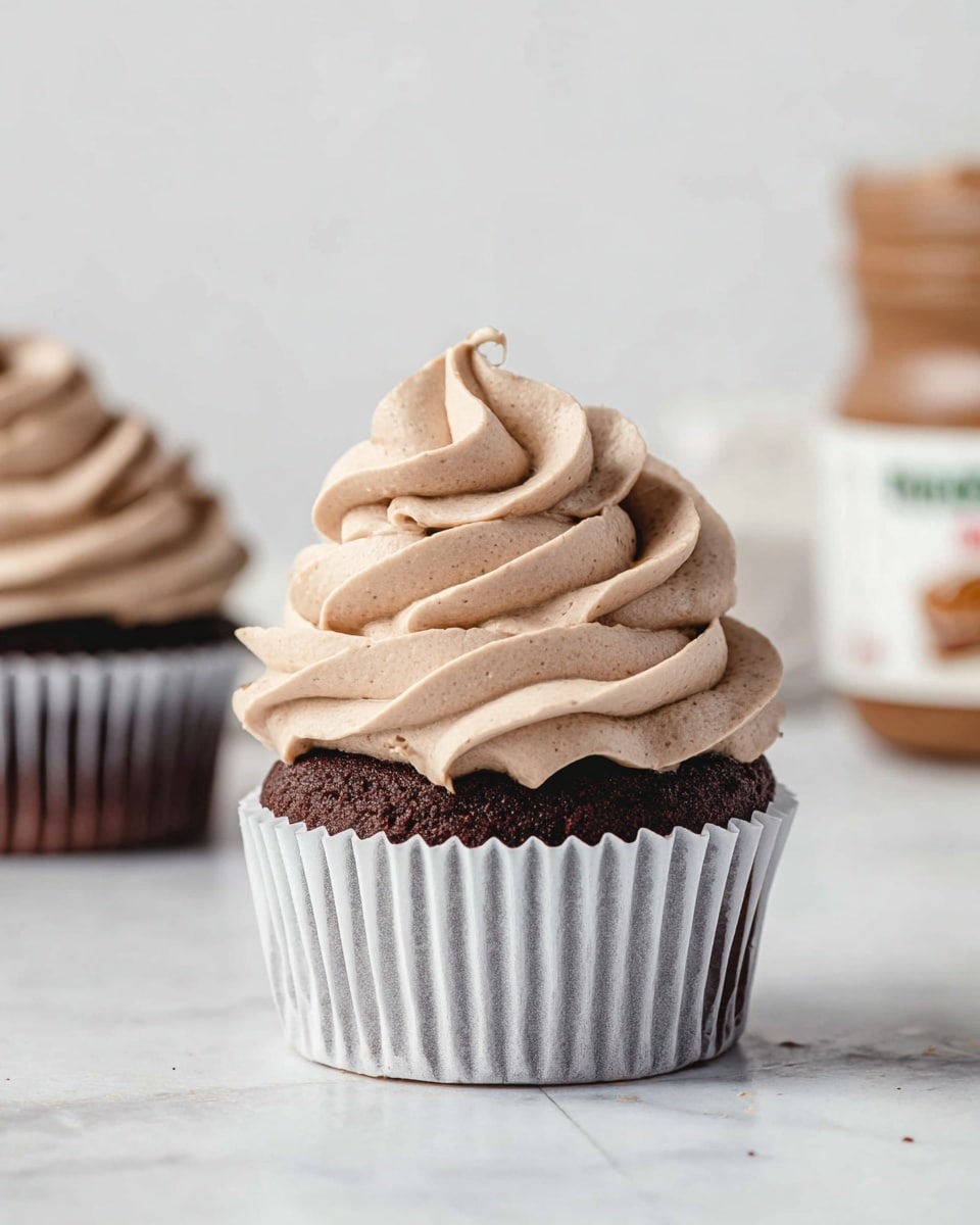 A chocolate cupcake sits centered with a thick swirl of light brown, creamy frosting on top, shaped in smooth, soft peaks and folds. The cake base is dark chocolate, visible through the white cupcake liner with vertical ridges, creating a contrast with the frosting. Part of a small jar, likely of hazelnut spread, is blurred in the background on the right side, while another cupcake with similar frosting sits slightly out of focus to the left. The entire scene rests on a white marbled textured surface with a plain white background. photo taken with an iphone --ar 4:5 --v 7