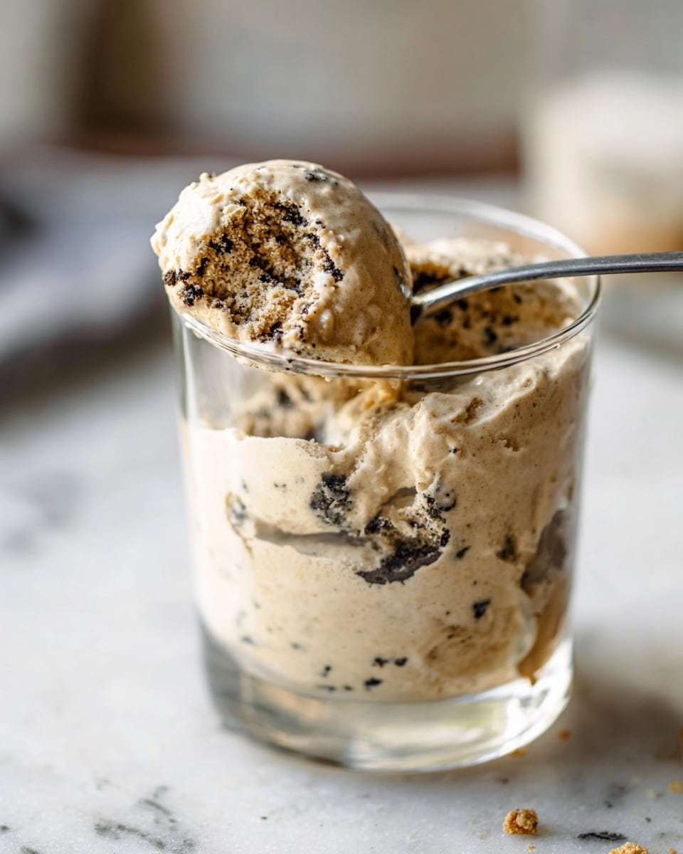 A close-up view of a clear glass cup filled with creamy, light beige ice cream mixed with crushed dark chocolate cookie pieces spread through the ice cream layer, topped with more cookie crumbs and chunks. A silver spoon is dipped into the ice cream, scooping up some, positioned on the left side of the cup. Below the cup on a white marbled surface, there are three dark chocolate sandwich cookies, one split in half showing the white cream filling. To the right of the cup, a black ice cream scoop rests on the surface, slightly out of focus. Photo taken with an iphone --ar 4:5 --v 7