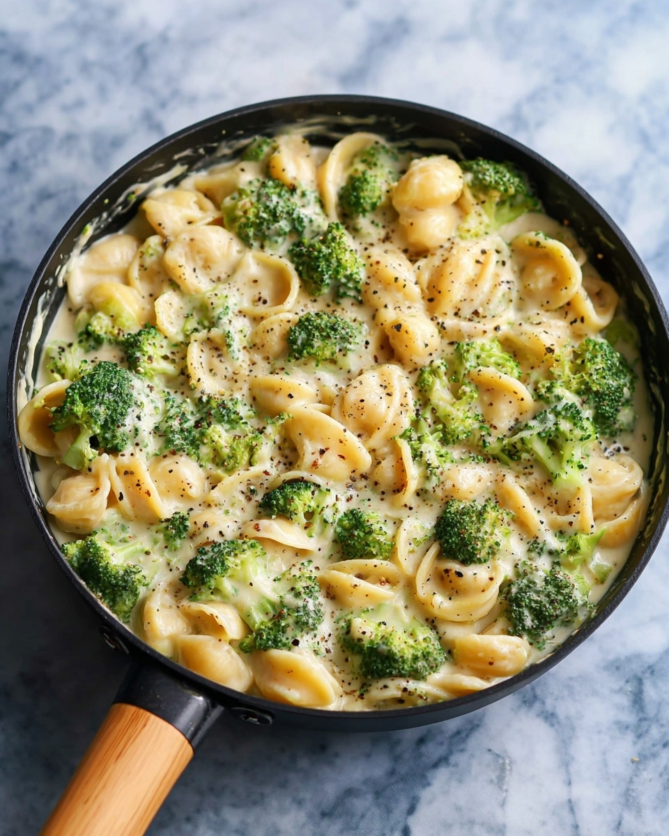 The image shows a black skillet with a wooden handle filled with creamy pasta and broccoli. The dish has three main visible layers: bright green broccoli florets scattered evenly throughout, yellow shell pasta pieces mixed well with the broccoli, and a smooth, thick, white cream sauce covering everything. Black pepper is sprinkled lightly on top, adding small dark specks around the dish. The skillet rests on a white marbled texture surface. Photo taken with an iphone --ar 4:5 --v 7