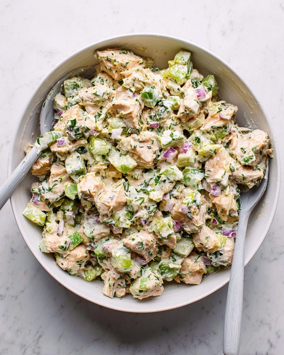A white bowl filled with a creamy salad made of chopped light orange pieces of chicken mixed with small green celery chunks, tiny purple bits of red onion, and finely chopped green herbs, all coated in a thick white mayo-like dressing. Two light gray spoons rest inside the bowl on opposite sides, one partially covered by the salad. The bowl sits on a white marbled surface. photo taken with an iphone --ar 4:5 --v 7