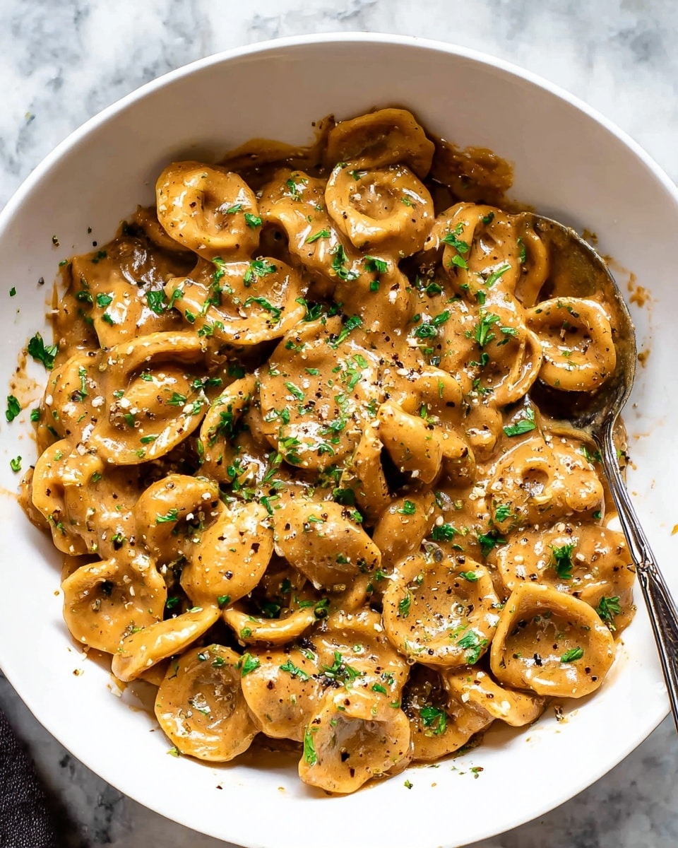 A white bowl filled with a creamy pasta dish made of orecchiette pasta coated in a thick, light brown sauce with visible black pepper specks. The pasta is garnished with finely chopped green herbs evenly spread on top. A silver spoon is partially immersed in the pasta on the right side inside the bowl. The bowl sits on a white marbled surface. Photo taken with an iphone --ar 4:5 --v 7