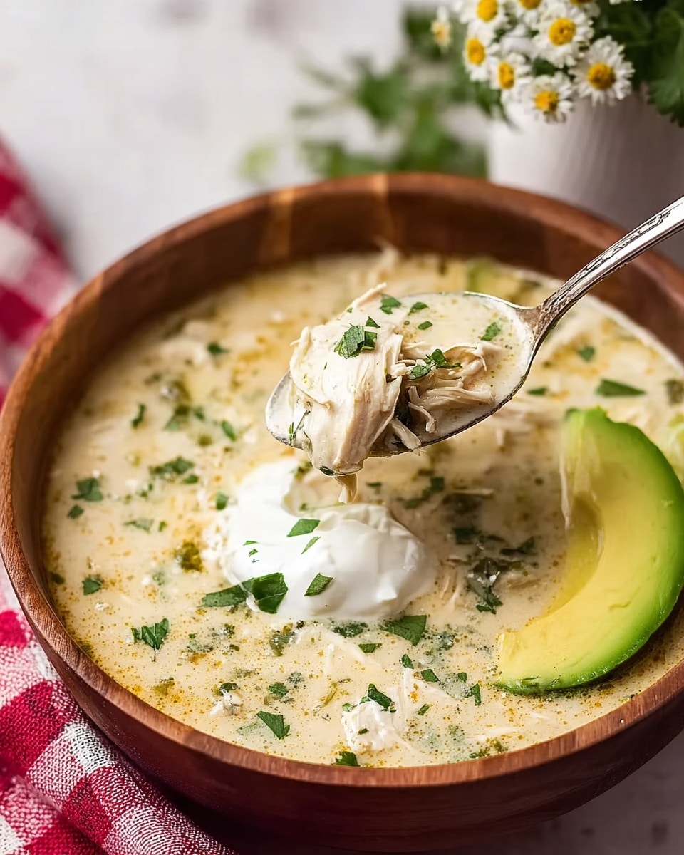 A wooden bowl filled with creamy light beige soup speckled with green herbs, with visible pieces of shredded white chicken mixed throughout. On the surface, there are dollops of white sour cream and a wedge of bright green avocado placed on the right side of the bowl. A silver spoon lifts a portion of the soup, showing chicken, sour cream, and herbs close up. The bowl sits on a white marbled surface, with a red and white checkered cloth partially visible beneath it, and delicate white flowers in a white vase in the soft blurred background. Photo taken with an iphone --ar 4:5 --v 7