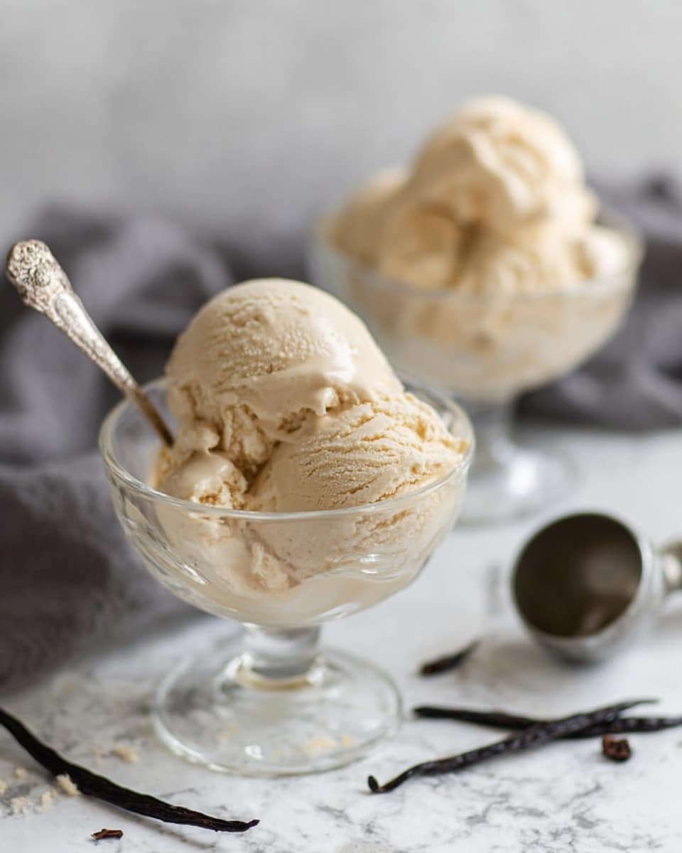 Two clear glass dessert cups filled with creamy light beige ice cream are placed on a white marbled surface. Each cup holds about two rounded scoops with a smooth, slightly textured surface. One cup is closer to the camera and has a small ornate silver spoon resting inside. In the background, the second cup appears slightly blurred. Scattered around the cups are small pieces of dark vanilla bean and an empty metal ice cream scoop lies to the right. A soft gray cloth adds gentle contrast in the back. Photo taken with an iphone --ar 4:5 --v 7