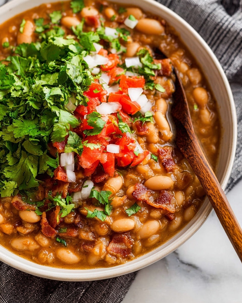 A close-up view shows a bowl filled with cooked white beans in a thick brown sauce. There are small pieces of red tomatoes, bits of green herbs, and chopped white onions mixed in the dish. On top, fresh green cilantro leaves add color. A wooden spoon rests inside the bowl on the right side. The bowl itself is dark gray and sits on a striped cloth on a white marbled surface. In the background, a sliced lime half and a whole green chili pepper sit beside the bowl. photo taken with an iphone --ar 4:5 --v 7