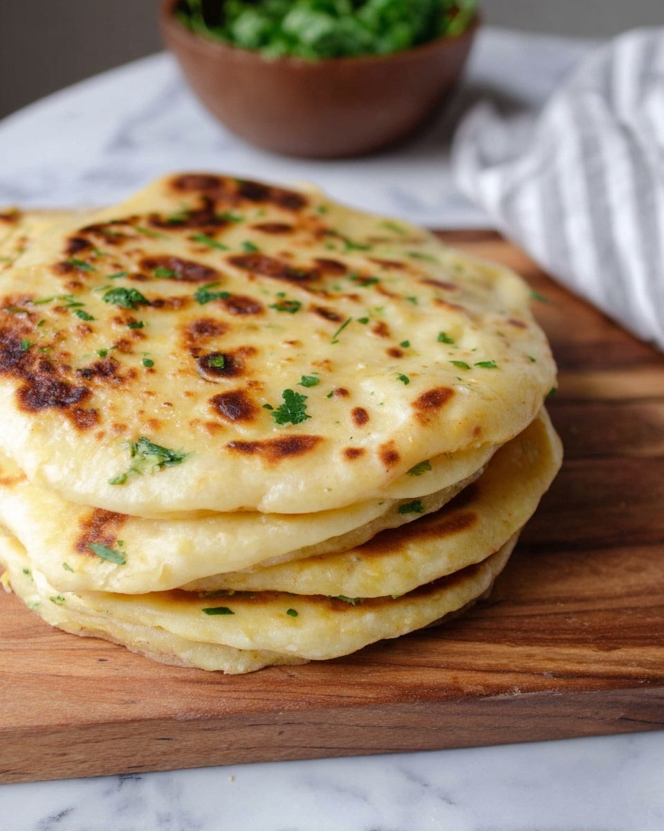A stack of five golden brown flatbreads is shown on a wooden cutting board resting on a white marbled surface. The top flatbread has a slightly crispy texture with browned spots and small green herb bits scattered across its surface. Each flatbread layer is thick and soft with irregular edges, slightly off-white inside with a light bready texture. In the background, a blurred brown bowl filled with fresh green herbs is visible. Photo taken with an iphone --ar 4:5 --v 7